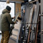Osoba porównuje narty w wypożyczalni przy stojaku ze sprzętem.
