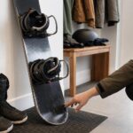 Osoba ogląda używany snowboard stojący przy ścianie, obok leżą buty snowboardowe.