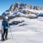 Osoba przy łagodnym stoku narciarskim w Dolomitach patrzy na mapę tras, w tle szerokie ośnieżone zbocza.