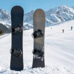 Dwie deski snowboardowe stoją w śniegu przy stoku, z rozmytym tłem górskiego ośrodka.