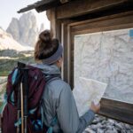 Osoba sprawdza mapę szlaku przy tablicy w Dolomitach, z górską ścieżką i szczytami w tle.
